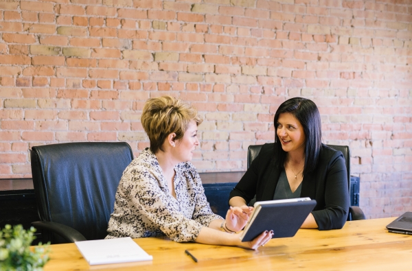 Two women have a conversation at a conference table in front of a brick wall.